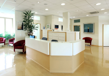 Heller, moderner Empfangsbereich in einem Büro oder einer Klinik. Es gibt eine zentrale Empfangstheke mit zwei Stühlen dahinter, Topfpflanze, Schränke im Hintergrund und mehrere rote Stühle im Wartebereich. Tageslicht kommt durch die Fenster herein.