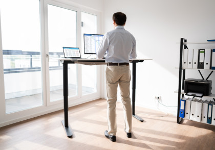 Ein Mann steht vor einem höhenverstellbaren Schreibtisch in einem modernen Büro. Auf dem Schreibtisch befinden sich ein Laptop und ein zusätzlicher Bildschirm mit Tabellen und Diagrammen.