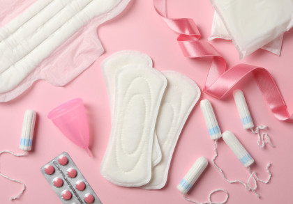 Das Bild zeigt verschiedene Menstruationsprodukte auf einem rosa Hintergrund. Zu sehen sind Damenbinden, Tampons, eine Menstruationstasse, Schmerztabletten und eine rosafarbene Schleife.