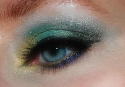 Nahaufnahme eines geschminkten Auges mit blau-grünem Lidschatten, schwarzen Eyeliner und Mascara. Teile des Unterlids sind dunkelblau und das innere Augenlid ist mit einem goldenen Lidschatten versehen.