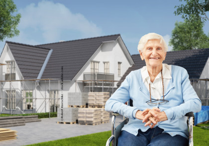Eine ältere Frau in einem blauen Pullover sitzt im Rollstuhl und lächelt. Im Hintergrund befindet sich ein im Bau befindliches modernes zweistöckiges Haus mit Gerüsten, Baumaterialien und Bäumen. Der Himmel ist klar und blau.