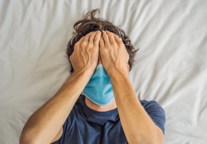 Eine Person liegt auf einem Bett mit einem weißen Laken und trägt eine blaue Gesichtsmaske. Die Person hält sich mit beiden Händen den Kopf und hat die Augen geschlossen, was auf Stress oder Müdigkeit hinweisen könnte.