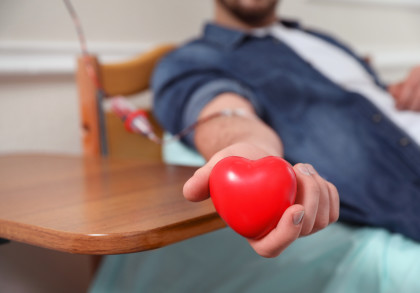 Ein Mann spendet Blut und liegt auf einem Stuhl. Er hält ein rotes Herz aus Gummi in seiner Hand. Ein Schlauch führt von seinem Arm zur Blutsammelvorrichtung.