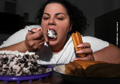 Eine Frau in einem weißen T-Shirt isst Kuchen mit einer Gabel, während sie in der anderen Hand einen Churro hält. Vor ihr auf dem Tisch befindet sich ein großer Stück Kuchen auf einem blauen Teller. Die Szene spielt sich in einer dunklen Küche ab.