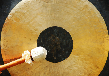 Nahaufnahme eines großen Gongs. Im Vordergrund wird ein Schlägel, umwickelt mit weißem Stoff, von einer Hand gehalten und ist bereit, den Gong zu schlagen. Der Gong hat ein dunkles Zentrum, das von einem goldenen Ring umgeben ist.