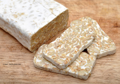 Ein Block Tempeh liegt auf einem Holzschneidebrett. Neben dem Block sind einige Scheiben Tempeh geschnitten und liegen ebenfalls auf dem Brett.