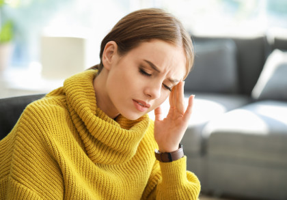 Eine Frau in einem gelben Strickpullover sitzt auf einem Sofa und hält sich den Kopf vor Schmerzen. Sie wirkt, als habe sie Kopfschmerzen. Im Hintergrund ist ein unscharfer Raum zu sehen.