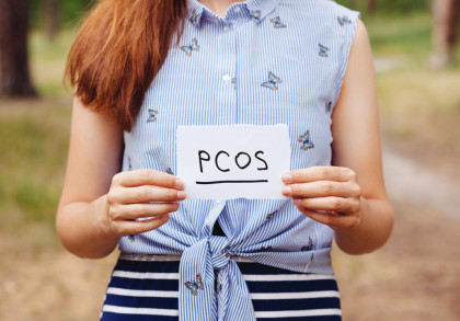 Eine Person hält ein handgeschriebenes Schild mit der Aufschrift "PCOS". Sie trägt ein ärmelloses, blau-weiß gestreiftes Hemd mit Schmetterlingsmustern und einen blau-weiß gestreiften Rock. Im Hintergrund ist eine verschwommene, grüne Outdoor-Umgebung.