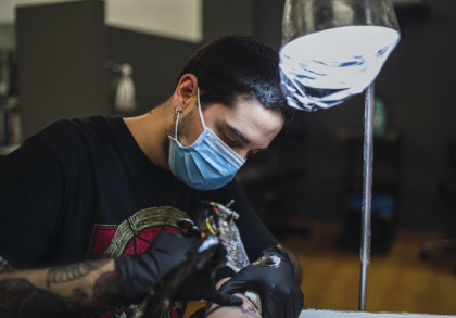 Ein Tätowierer mit schwarzem T-Shirt und Einweghandschuhen tätowiert den Arm einer Person. Er trägt eine blaue Gesichtsmaske und konzentriert sich auf seine Arbeit, während eine Lampe über ihm leuchtet.