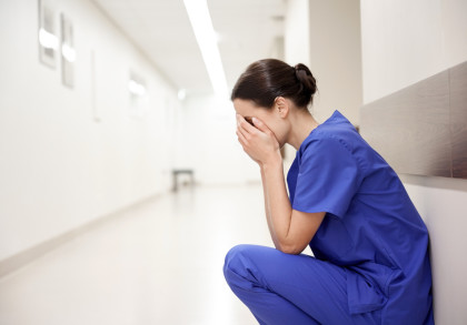 Eine Krankenschwester in blauer medizinischer Kleidung sitzt auf dem Boden eines hellen Flurs und hält sich traurig oder erschöpft das Gesicht mit den Händen.