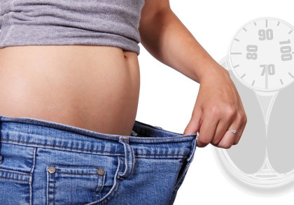 Eine Person trägt eine locker sitzende Jeans und hält sie am Bund fest. Der Bauch der Person ist sichtbar. Im Hintergrund ist eine Waage mit Kilogramm-Anzeigen zu sehen.