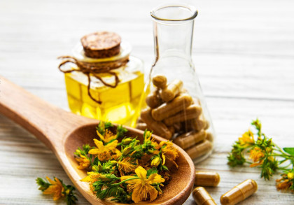Ein Holzlöffel mit gelben Blüten und grünen Blättern liegt auf einer weißen Holzoberfläche. Im Hintergrund steht eine kleine Glasflasche mit Korkverschluss, gefüllt mit einer gelben Flüssigkeit, sowie ein weiteres Glas mit Kapseln.