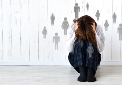 Eine Person sitzt zusammengekauert auf dem Boden, mit dem Kopf in den Händen, offensichtlich gestresst oder verzweifelt. Im Hintergrund sind unscharfe Silhouetten von Personen zu sehen, die ein Gefühl der Überwältigung oder des Drucks darstellen könnten.
