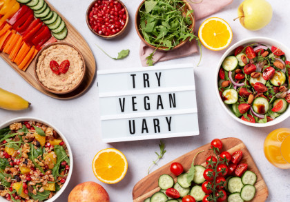 Ein Tisch gedeckt mit verschiedenen veganen Lebensmitteln: Paprika, Gurken, Karotten, Humus, Granatapfelkerne, Rucola, eine Orangenscheibe, ein Apfel, ein Glas Orangensaft, eine Banane und Salate. Dazu ein Schild mit der Aufschrift "Try Veganuary".