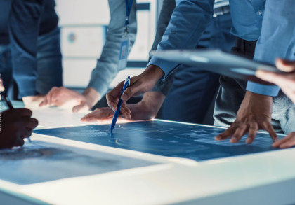 Mehrere Personen beugen sich über einen beleuchteten Tisch und arbeiten gemeinsam an Plänen oder technischen Zeichnungen.