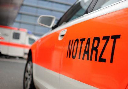 Ein orange-weißes Notarztfahrzeug mit der Aufschrift "NOTARZT" auf der Seite ist in Nahaufnahme zu sehen. Im Hintergrund befindet sich ein weiterer Krankenwagen vor einem modernen Gebäude.