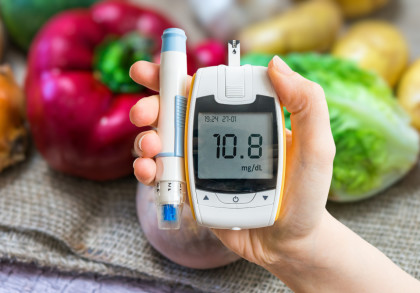 Eine Hand hält ein Blutzuckermessgerät, das einen Wert von 70,8 mg/dL anzeigt. Neben dem Blutzuckermessgerät wird ein Stechhilfe-Instrument gehalten. Im Hintergrund sind verschiedene Gemüsesorten wie ein roter Paprika und grüner Salat zu sehen.