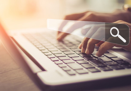 Eine Person tippt auf einer Laptop-Tastatur, während sie eine virtuelle Suchleiste mit einer Lupe verwendet. Das Bild ist in warmen, gedämpften Farben gehalten.