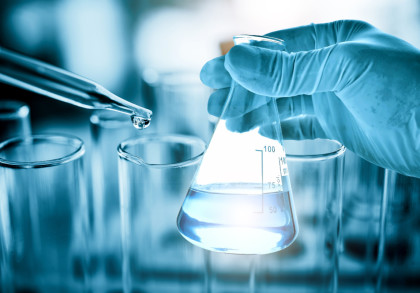 Nahaufnahme einer Hand mit blauen Handschuhen, die ein Erlenmeyerkolben mit einer klaren bläulichen Flüssigkeit hält. Eine Pipette lässt einen Tropfen Flüssigkeit in ein Reagenzglas fallen. Im Hintergrund mehrere unscharfe Reagenzgläser.
