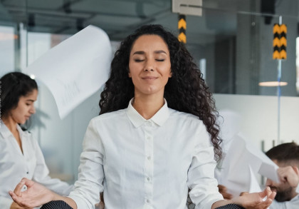Eine Frau mit lockigem Haar und einem weißen Hemd sitzt in einem Büro und meditiert, während sie die Augen geschlossen hat. Im Hintergrund sind andere Personen zu sehen, von denen einige Dokumente in die Luft werfen.