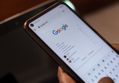 Nahaufnahme eines Smartphones, auf dem die Google-Suchseite geöffnet ist. Die Person gibt gerade eine Suchanfrage ein. Man sieht das Google-Logo oben auf dem Bildschirm und einige Suchvorschläge darunter. Der Hintergrund ist verschwommen.