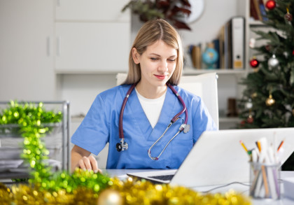 Ärztin in OP-Kleidung mit Stethoskop sitzt in einem weihnachtlich geschmückten Büro vor dem Computer.