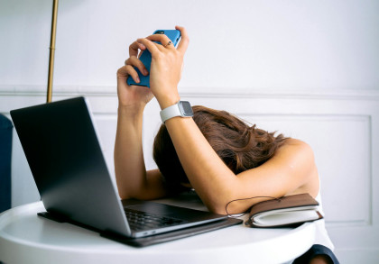 Eine Frau, die ihren Kopf auf Ihrem PC-Tisch ablegt und die Arme über den Kopf zusammenschlägt. In ihren Händen befindet sich ein Handy. 