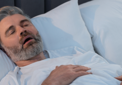 Mittelalter Mann mit grauem Bart schläft in Rückenlage im Bett mit geöffnetem Mund, Hinweise auf mögliche Schlafapnoe oder Schnarchen. Schlafzimmerumgebung mit weißen Kissen und Bettdecke, Fokus auf Atemprobleme im Schlaf.