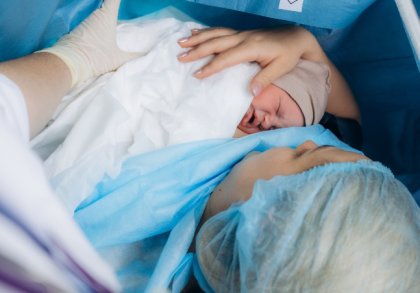 Frau liegt während eines Kaiserschnitts im Operationssaal, hält ihr Neugeborenes zum ersten Hautkontakt im Arm. Das Baby wird von medizinischem Personal unterstützt und liegt geschützt in Tüchern.