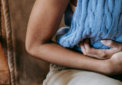 Person hält sich mit einem blauen Wärmekissen den Bauch, sitzt leicht nach vorne gebeugt und zeigt sichtbare Bauchschmerzen – symbolische Darstellung von krampfartigen Beschwerden wie Koliken oder starken Unterbauchschmerzen.