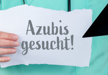 Eine Person im grünen Kasak mit Stethoskop um den Hals hält ein Blatt auf dem "Azubis gesucht" steht. In der anderen Hand hält sie einen schwarzen Pfeil, der auf das Blatt zeigt.