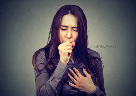 Eine Frau mit langen, braunen Haaren trägt eine graue Bluse. Sie hat die Augen geschlossen und hält sich die Hand vor den Mund, als ob sie hustet. Mit der anderen Hand hält sie sich die Brust, was auf Unwohlsein oder Schmerzen hinweist.