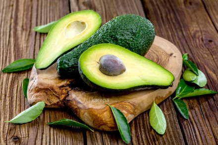 Zwei Hälften und eine ganze Avocado liegen auf einem hölzernen Schneidebrett, umgeben von grünen Blättern auf einem Holztisch. Die eine Hälfte zeigt den runden Kern, während die andere Hälfte entkernt ist.