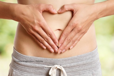 Nahaufnahme eines nackten Bauches mit Händen, die eine herzförmige Geste machen. Die Person trägt graue Freizeithosen. Im Hintergrund ist ein unscharfer, natürlicher Hintergrund zu sehen.