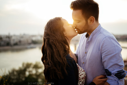 Ein Paar steht bei Sonnenuntergang am Wasser und küsst sich innig. Der Mann trägt ein hellblaues Hemd, die Frau hat langes, braunes Haar. Im Hintergrund ist eine unscharfe Stadtlandschaft zu sehen.