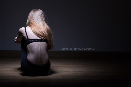Eine Frau mit langen, blonden Haaren sitzt auf dem Boden mit dem Rücken zur Kamera. Sie trägt ein schwarzes Oberteil. Die Szene ist dunkel mit einem Spotlicht, das den Mittelteil ihres Rückens hervorhebt.