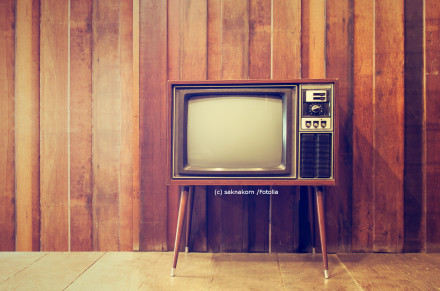 Ein retro Fernseher mit Holzgehäuse und vier Beinen steht vor einer Wand mit holzverkleideten Paneelen. Der Bildschirm des Fernsehers ist ausgeschaltet.