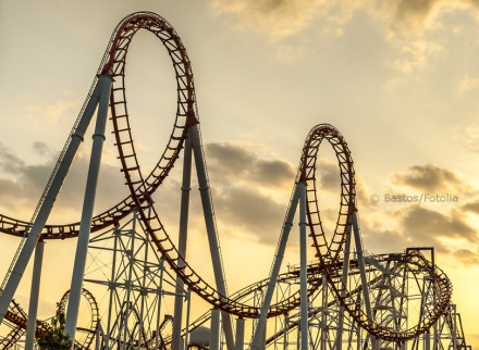 Das Bild zeigt eine Achterbahn mit mehreren Loopings und hohen Stützen vor einem Himmel bei Sonnenuntergang. Die Struktur der Achterbahn hebt sich deutlich gegen den orangefarbenen Himmel mit vereinzelten Wolken ab.