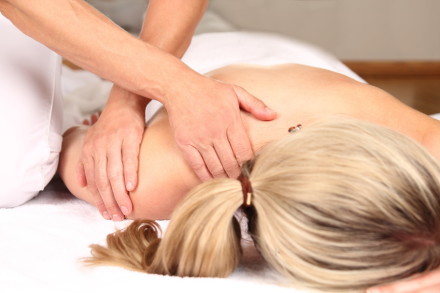 Eine Person, die auf einem Massagetisch liegt, erhält eine Rückenmassage. Eine andere Person massiert mit beiden Händen den Rücken der liegenden Person. Die liegende Person hat blonde Haare, die im Nacken zu einem Zopf gebunden sind.