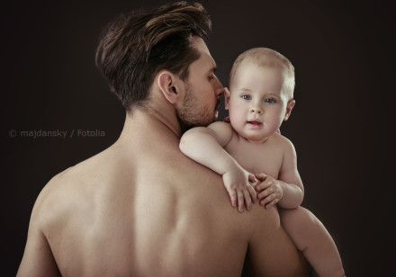 Ein Mann hält ein Baby auf dem Arm. Beide sind oberkörperfrei. Der Mann ist von hinten zu sehen, während das Baby nach vorne schaut. Der Hintergrund ist dunkel und schlicht.