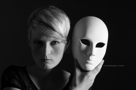 Schwarz-Weiß-Foto einer Person mit kurzem, hellem Haar, die eine weiße Maske neben ihrem Gesicht hält. Die Person schaut direkt in die Kamera, und ein starker Schatten fällt auf die Hälfte ihres Gesichts.