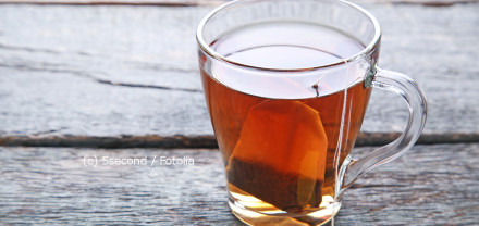 Eine Glas-Tasse mit heißem Tee und einem Teebeutel darin steht auf einer rustikalen, grauen Holzoberfläche.