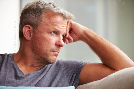 Ein nachdenklicher Mann mit grauen Haaren sitzt auf einem Sofa und stützt den Kopf auf seine Hand. Er trägt ein graues T-Shirt und schaut aus dem Fenster.