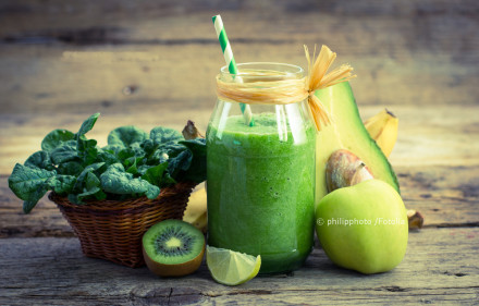 Grüner Smoothie in einem Glas mit Strohhalm, umgeben von frischem Spinat in einem Korb, einer halben Kiwi, einer Zitronenscheibe, einem Apfel und einer Avocado-Hälfte, die auf einem rustikalen Holzuntergrund arrangiert sind.