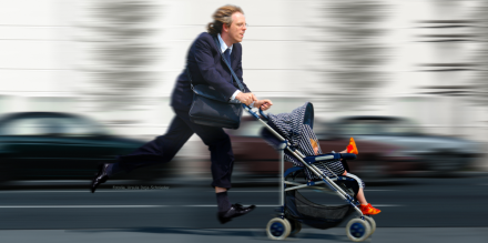 Ein Mann im Anzug läuft eilig mit einem Kinderwagen über eine Straße. Er scheint in Eile zu sein, während er ein Kind im Wagen schiebt. Die Umgebung ist verschwommen, was auf die Geschwindigkeit hinweist.