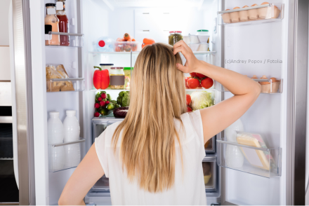 Eine Frau mit langen blonden Haaren steht vor einem offenen Kühlschrank, der mit verschiedenen Lebensmitteln gefüllt ist. Sie kratzt sich am Kopf und scheint darüber nachzudenken, was sie nehmen soll.