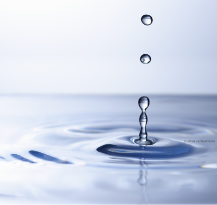 Tropfende Wassertropfen, die auf eine Wasseroberfläche fallen und konzentrische Kreise bilden. Die Oberfläche ist glatt und reflektierend, und der Hintergrund ist in weichen Blau- und Grautönen gehalten.