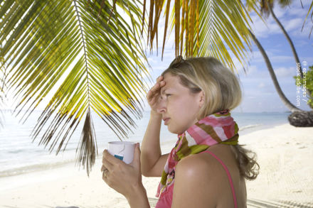 Eine Frau steht am Strand unter Palmen. Sie trägt ein ärmelloses Oberteil und einen bunten Schal. In ihrer rechten Hand hält sie eine Tasse und mit der linken Hand stützt sie ihre Stirn. Im Hintergrund ist der Ozean und ein klarer Himmel zu sehen.