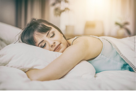Eine Frau schläft friedlich auf einem Bett mit weißer Bettwäsche. Sie liegt auf der Seite und ihr Kopf ruht auf einem weißen Kissen. Die Szene ist von weichem Licht durchflutet und im Hintergrund ist eine unscharfe Zimmeransicht zu sehen.