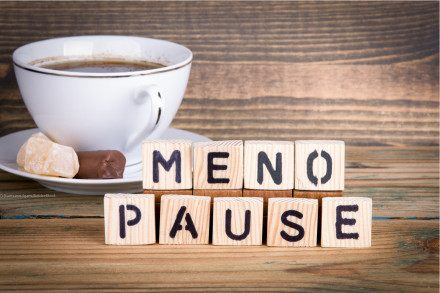 Eine weiße Tasse Kaffee mit einem kleinen Stück Schokolade und einem kleinen Gebäckstück auf einer Untertasse. Vor der Tasse befinden sich Holzklötze mit Buchstaben, die das Wort "MENOPAUSE" bilden.
